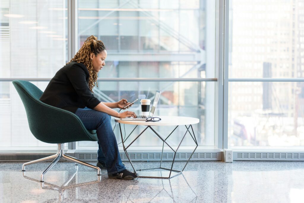 Imagem de uma mulher de perfil sentada em frente a um notebook, com um celular na mão.
Imagem: Pexels.