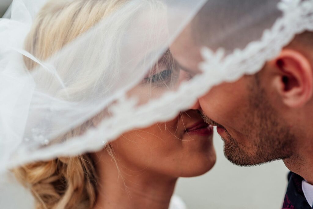 Imagem de um casal de noivos se beijando sob o véu da noiva. Imagem: Pexels.